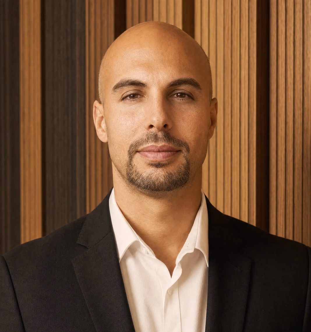 Shahar Kats, in a dark suit and open-collared white shirt, in front of a slatted wooden wall.