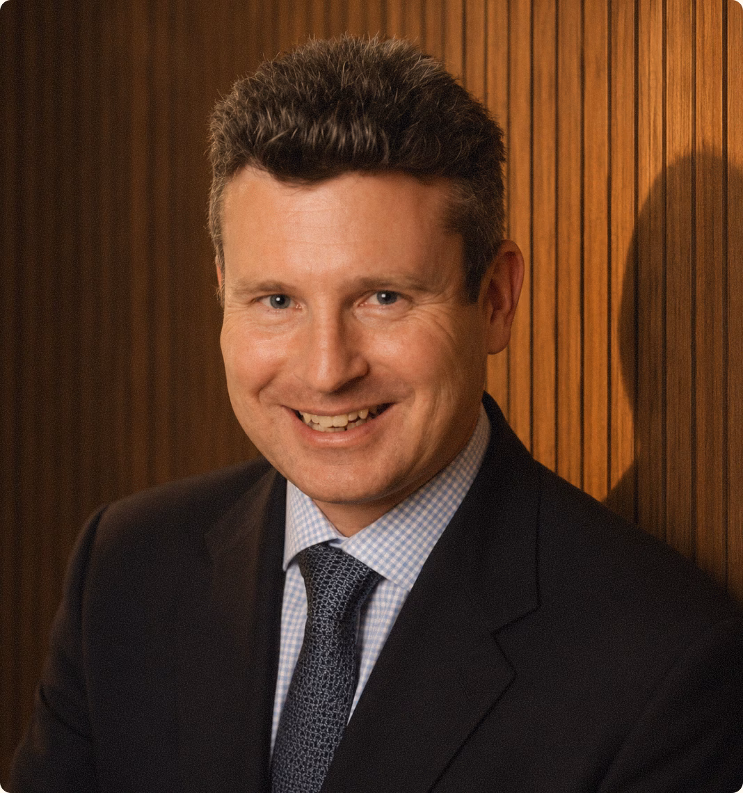 Euan Campbell, smiling, in a dark suit, checked shirt and patterned tie, in front of a slatted wooden wall.